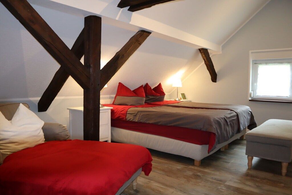 Ein Schlafzimmer mit schrägem Dach, Holzbalken, einem Bett mit roter Bettwäsche, einem Nachttisch und einem Fenster.