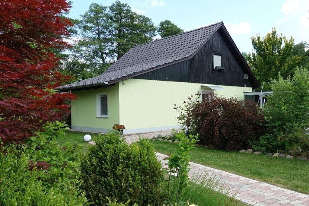 Een gelijkvloers huis met een schuin dak, een klein portaaltje en een verzorgde tuin met verschillende struiken en gazon.