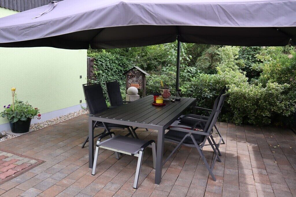 Un patio al aire libre con una mesa y sillas bajo un paraguas grande, rodeado de follaje.