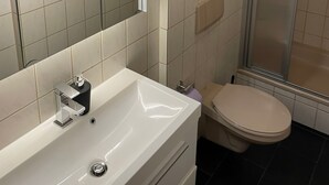 Kylpyhuone, jossa on valkoinen pesuallas, peili, wc-istuin ja lasinen suihkuseinä.