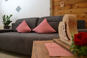 Un canapé gris avec des coussins rouges, une table en bois recouverte d’une nappe à carreaux, un panneau mural en bois et une plante en pot.