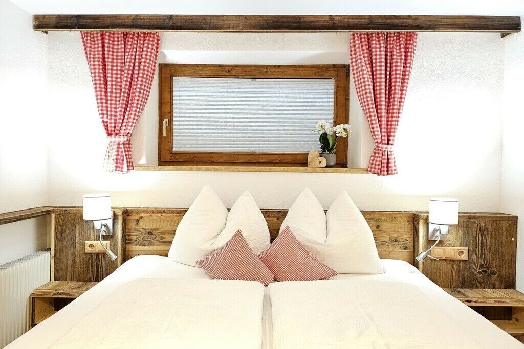 A double bed with a wooden headboard, two bedside tables with lamps, a window with checkered curtains, and a small shelf with a plant.
