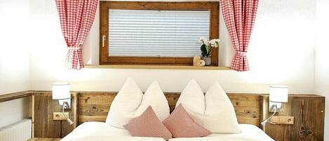 A double bed with a wooden headboard, two bedside tables with lamps, a window with checkered curtains, and a small shelf with a plant.
