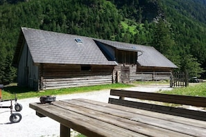 A rustic wooden building with a sloped roof, a bench in the foreground, and a forested mountain backdrop. - Self-catering hut Potzalm (Kleinsölk)