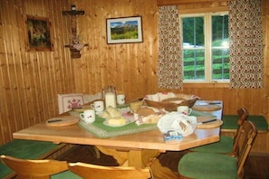 A wooden dining table with green cushioned chairs, a window with patterned curtains, and a framed picture on the wall. - Self-catering hut Potzalm (Kleinsölk)