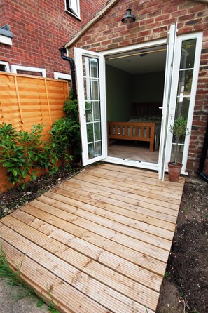 A wooden deck leading to a room with a bed, a potted plant, and a brick wall.