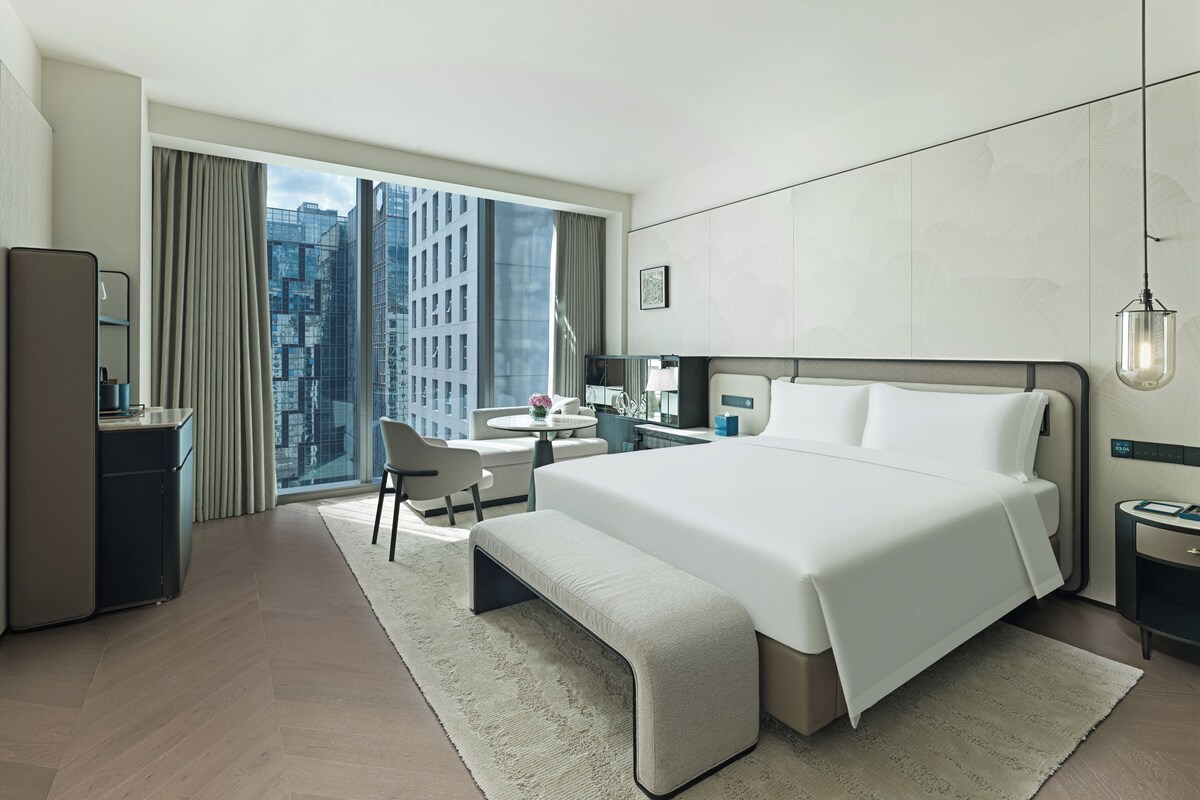 Quarto de hotel moderno com uma cama grande, uma escrivaninha, uma cadeira e vista para a paisagem urbana pela janela.