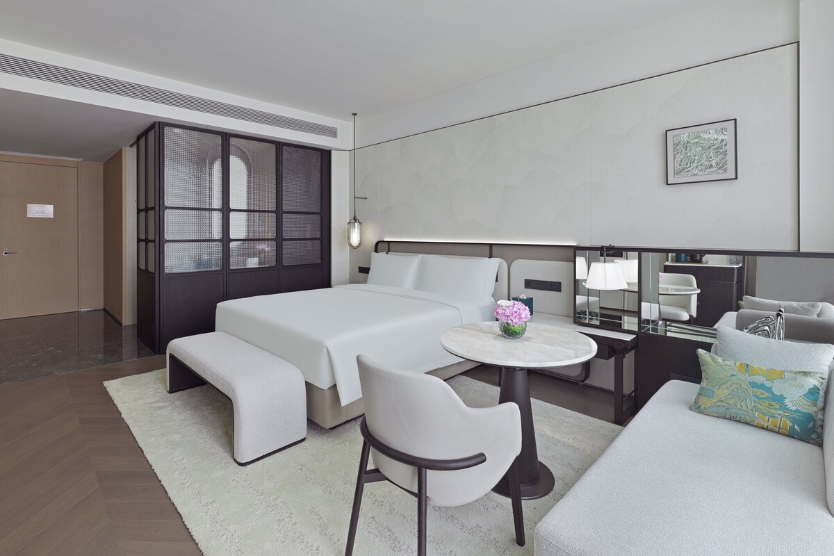 Quarto de hotel moderno com uma cama grande, área de estar com uma mesinha e cadeira, escrivaninha com luminária e guarda-roupa.