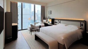 Ein modernes Hotelzimmer mit einem großen Bett, einem Schreibtisch, einem Sessel und Blick auf die Stadt durch die Fenster.