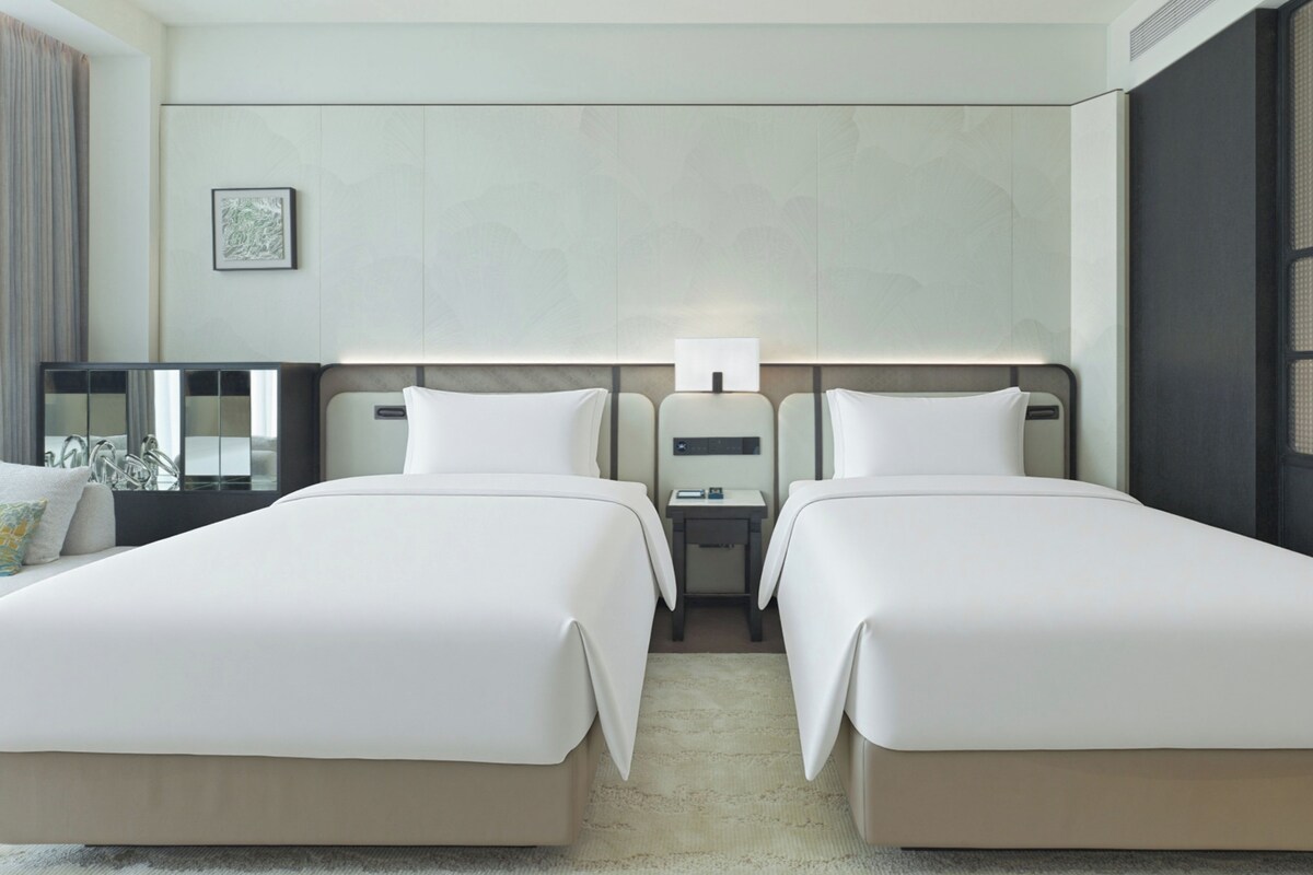 Quarto de hotel com duas camas, uma mesa de cabeceira com luminária e uma janela grande.