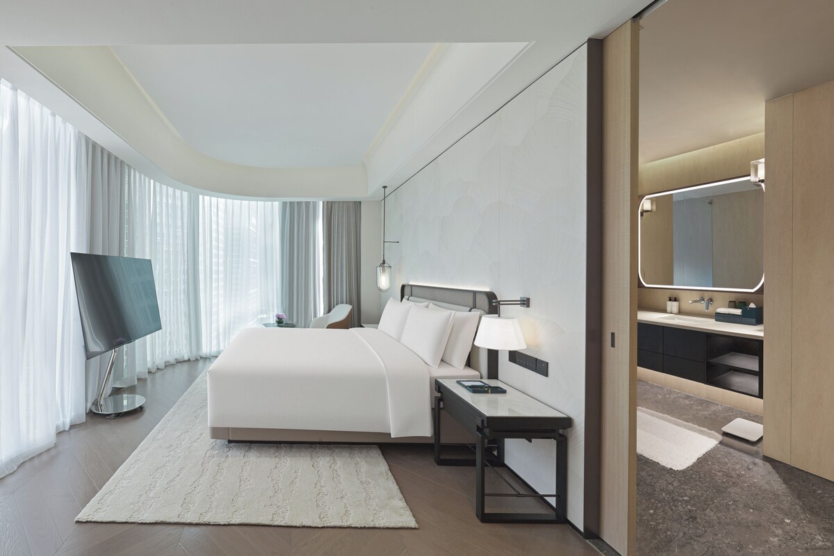 Quarto de hotel moderno com uma cama grande, televisão de tela plana, mesa de cabeceira, escrivaninha e banheiro com espelho e pia.
