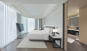 Quarto de hotel moderno com uma cama grande, televisão de tela plana, mesa de cabeceira, escrivaninha e banheiro com espelho e pia.