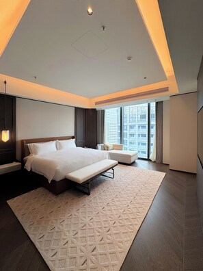 Quarto de hotel moderno com uma cama grande, um banco, área de estar e vista para a paisagem urbana.