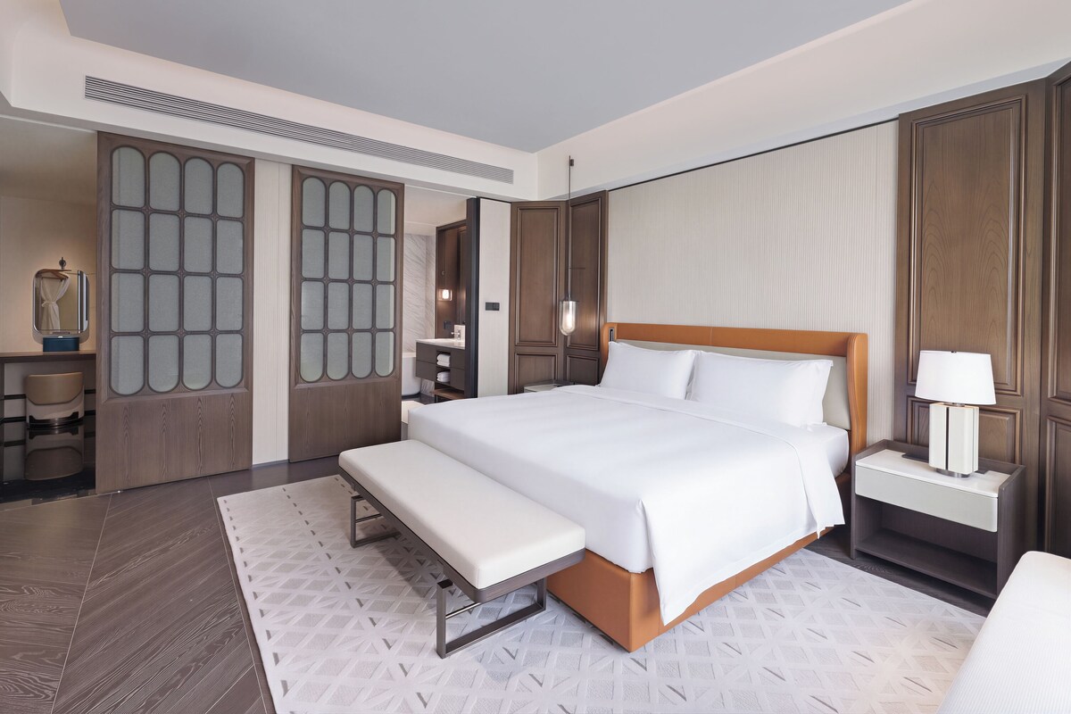 Quarto de hotel com uma cama grande, mesas de cabeceira, um banquinho, guarda-roupa e um espelho.
