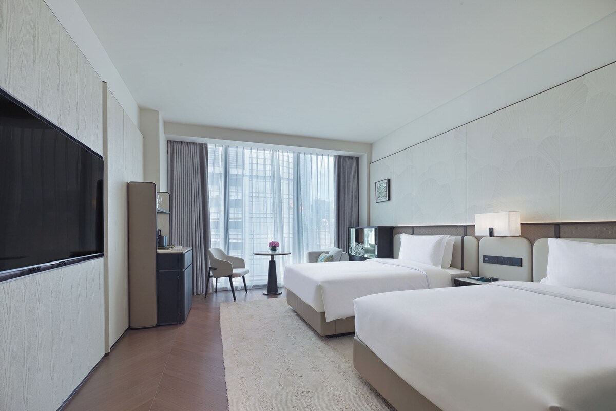 Quarto de hotel com duas camas, uma escrivaninha, uma cadeira, uma televisão e uma janela com cortinas.