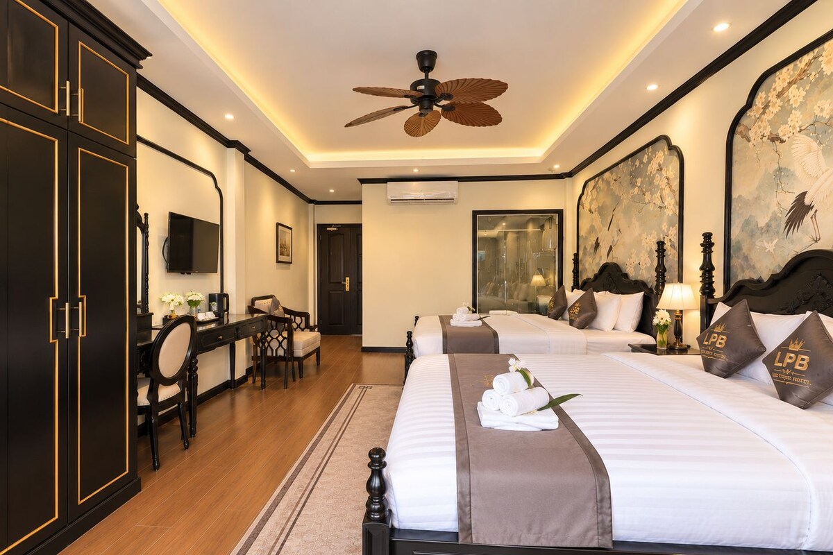 A hotel room with a large bed, a desk, a chair, a ceiling fan, and a large painting on the wall.