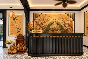 A reception desk with a 'RECEPTION' sign, surrounded by traditional decor including a vase with flowers, a fruit bowl, and a decorative stand. - Luang Prabang Indochine Heritage Hotel (Luang Prabang)