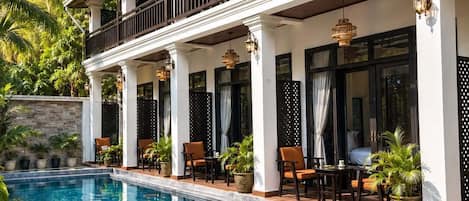 Zona junto a la piscina con piscina cristalina, mobiliario de exterior y un edificio con columnas y balcón.