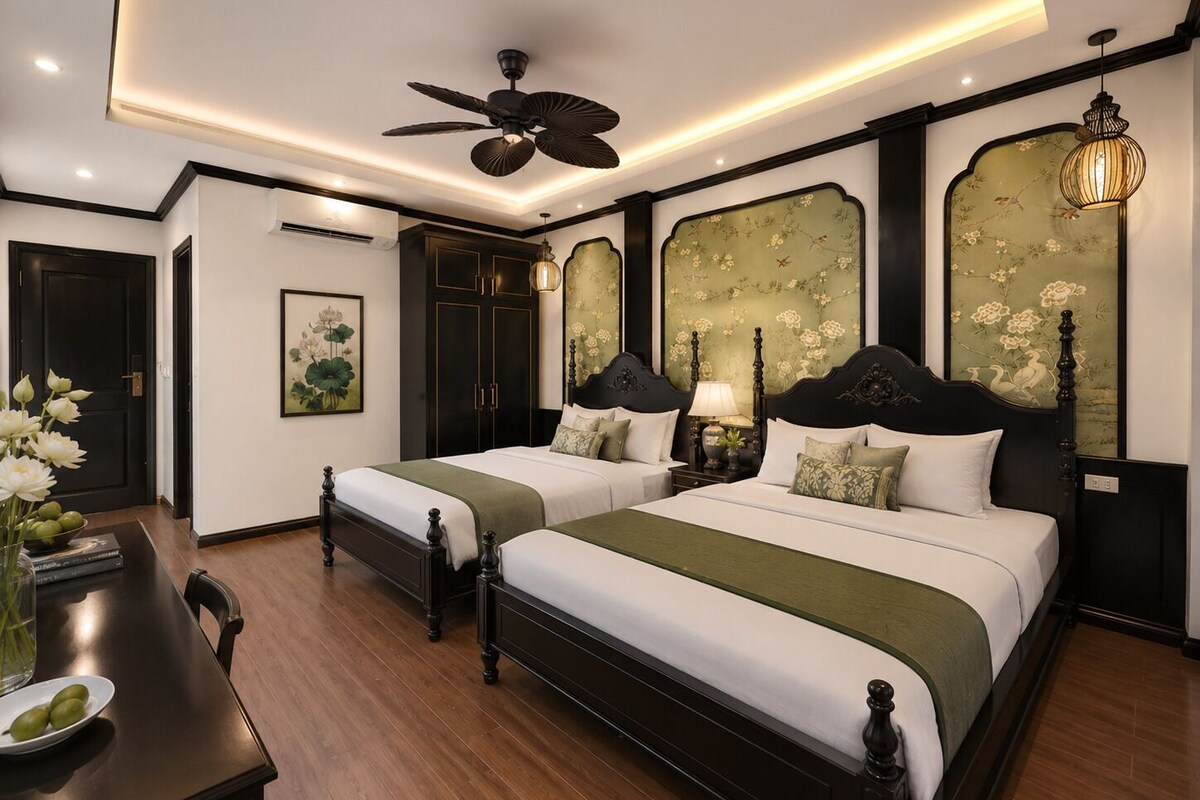 A hotel room with two beds, a desk, and a chair. There is a ceiling fan, a wall-mounted lamp, and a painting on the wall.