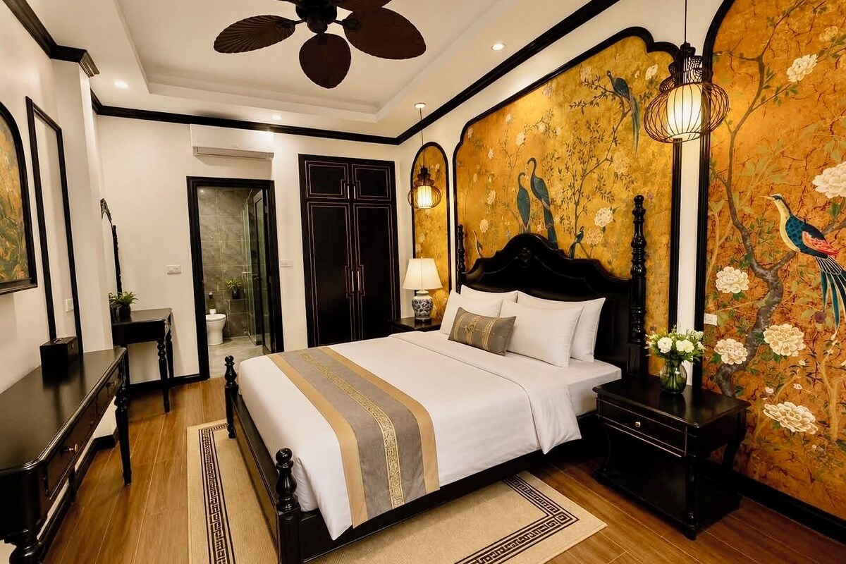 A hotel room with a large bed, a wooden floor, a ceiling fan, and a decorative wall with floral and bird motifs.
