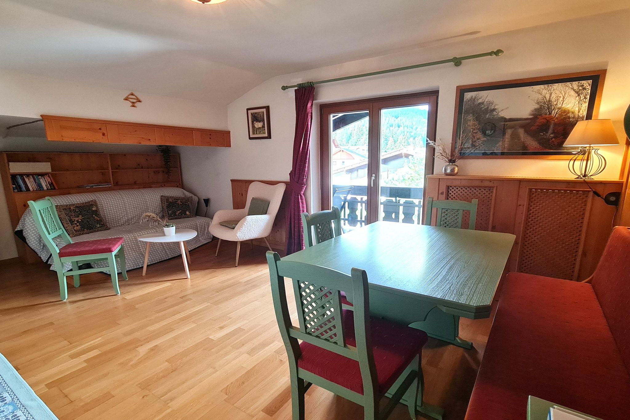 A cozy living room with a wooden floor, a sofa, a dining table, and chairs. There is a window with curtains, a bookshelf, and a painting on the wall.