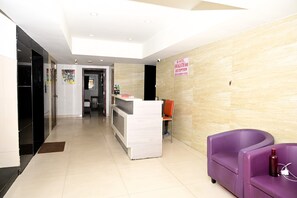 Zona de recepción de un hotel, con un mostrador, un área de descanso morada y un pasillo que conduce a otras habitaciones.