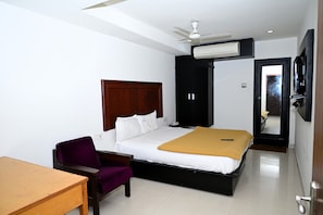 Habitación de hotel con cama, escritorio, silla, ventilador de techo, unidad de aire acondicionado y televisor.