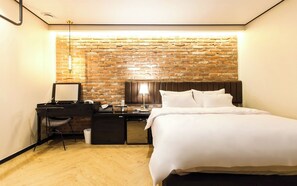 Una habitación de hotel con pared de ladrillo, una cama con ropa de cama blanca, un escritorio con una silla y una mesita de noche con una lámpara.