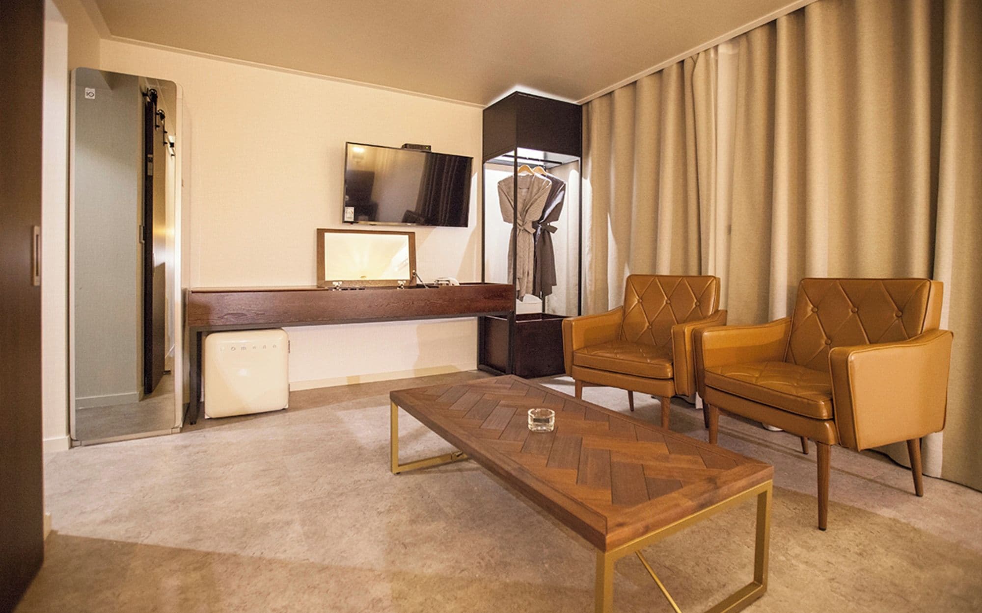 A hotel room with a wooden coffee table, two leather armchairs, a flat-screen TV, a desk with a computer, and a large window with curtains.