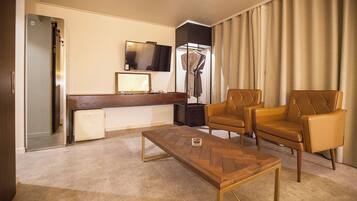 A hotel room with a wooden coffee table, two leather armchairs, a flat-screen TV, a desk with a computer, and a large window with curtains.