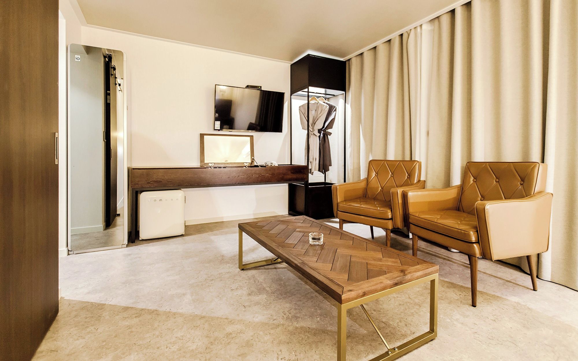 A hotel room with a wooden coffee table, two leather armchairs, a small desk with a mirror, and a wardrobe.