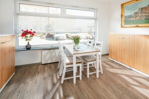 A modern kitchen with a dining table, white chairs, a window with blinds, and a painting on the wall. - Schmalenbeck House Lucius 1 (Norderney)