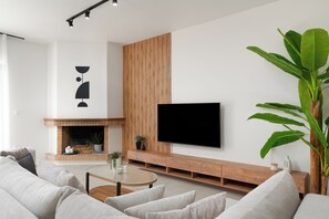 A modern living room with a fireplace, a flat-screen TV mounted on a wooden wall, and a large green plant. - Luxury 4Bedrooms Stay in North Athens (Agia Paraskevi)