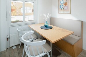 Een klein zithoekje met een houten tafel en twee witte stoelen, een vaas met bloemen, een radiator en een raam met uitzicht op gebouwen.