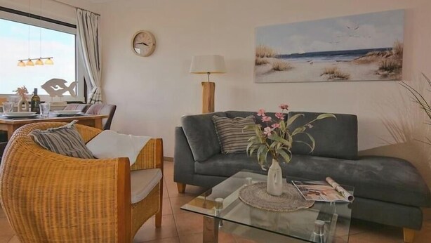 A living room with a sofa, a wicker chair, a glass coffee table, and a painting of a beach scene.