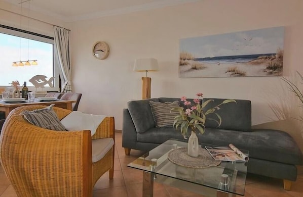 Un salon comprenant un canapé, un fauteuil en osier, une table basse en verre et un tableau représentant une scène de plage.