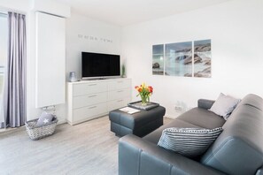 A modern living room with a flat-screen TV, a grey sofa, a coffee table with a vase of flowers, and a wall-mounted artwork. - Holiday apartment in Norderney with sauna (Norderney)