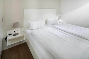 Ein sauberes, weißes Hotelzimmer mit einem Einzelbett, einem Nachttisch mit Lampe und einer Steckdose.