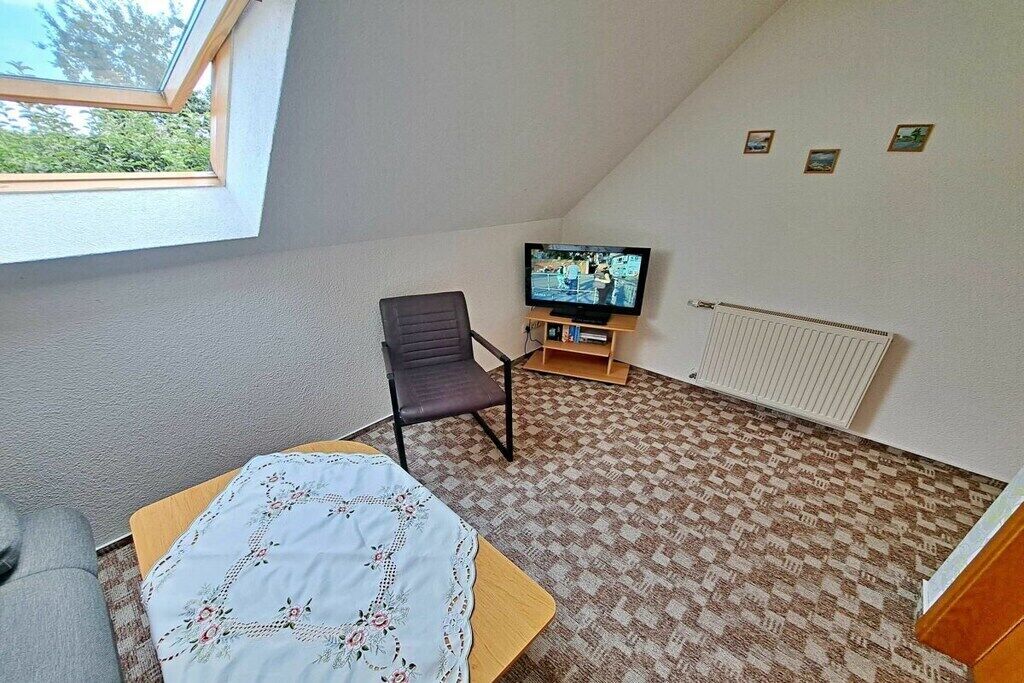 A room with a sloped ceiling, a television on a stand, a chair, a table with a tablecloth, and a patterned carpet.