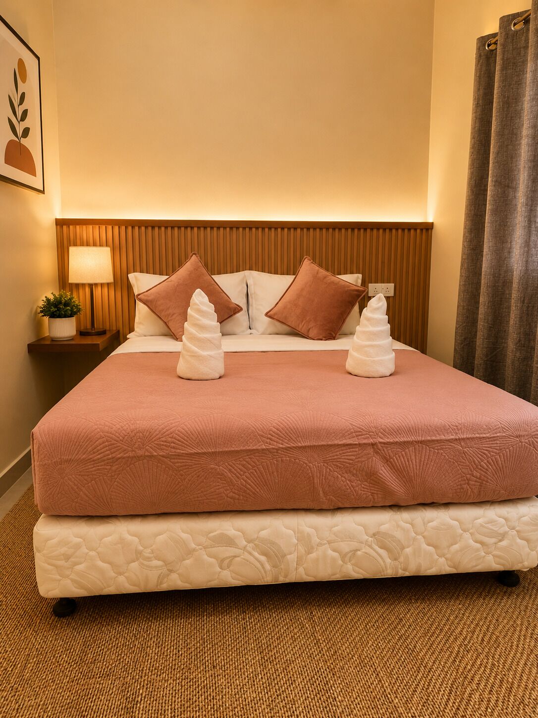 A hotel room with a bed, two pillows, a bedside lamp, a small plant, and a wall-mounted artwork.