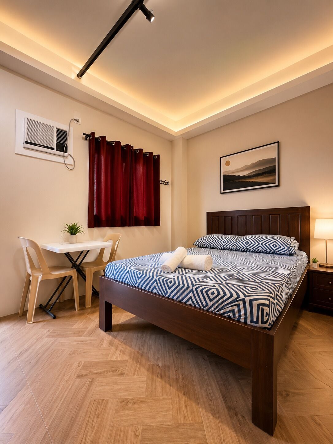 A bedroom with a bed, a desk, a chair, a window with curtains, an air conditioning unit, and a wall-mounted picture.
