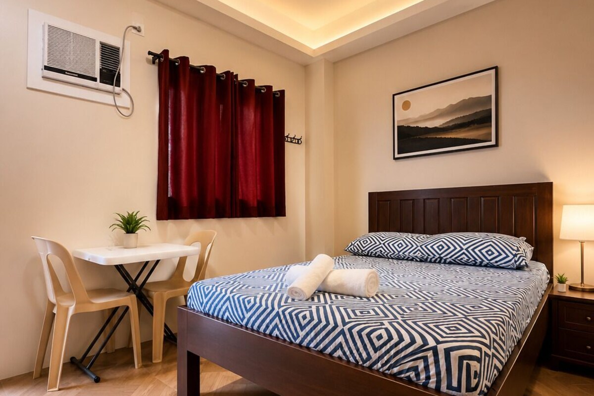 A bedroom with a bed, a desk, a chair, a window with curtains, an air conditioning unit, and a wall-mounted picture.