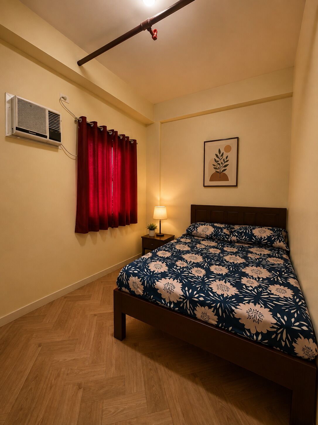 A bedroom with a wooden floor, a bed with a floral bedspread, a red curtain, a wall-mounted air conditioner, a bedside lamp, and a framed picture on the wall.