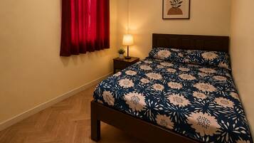 A bedroom with a wooden floor, a bed with a floral bedspread, a red curtain, a wall-mounted air conditioner, a bedside lamp, and a framed picture on the wall.