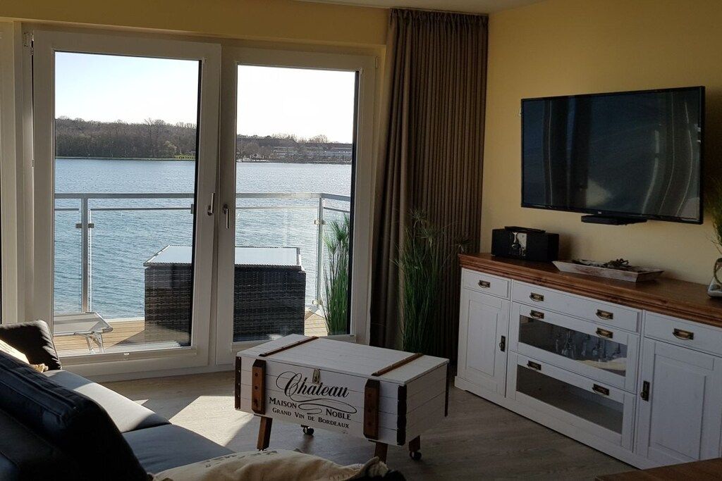A room with a sliding glass door leading to a balcony with a view of a lake. There is a television mounted on the wall and a wooden cabinet with drawers.