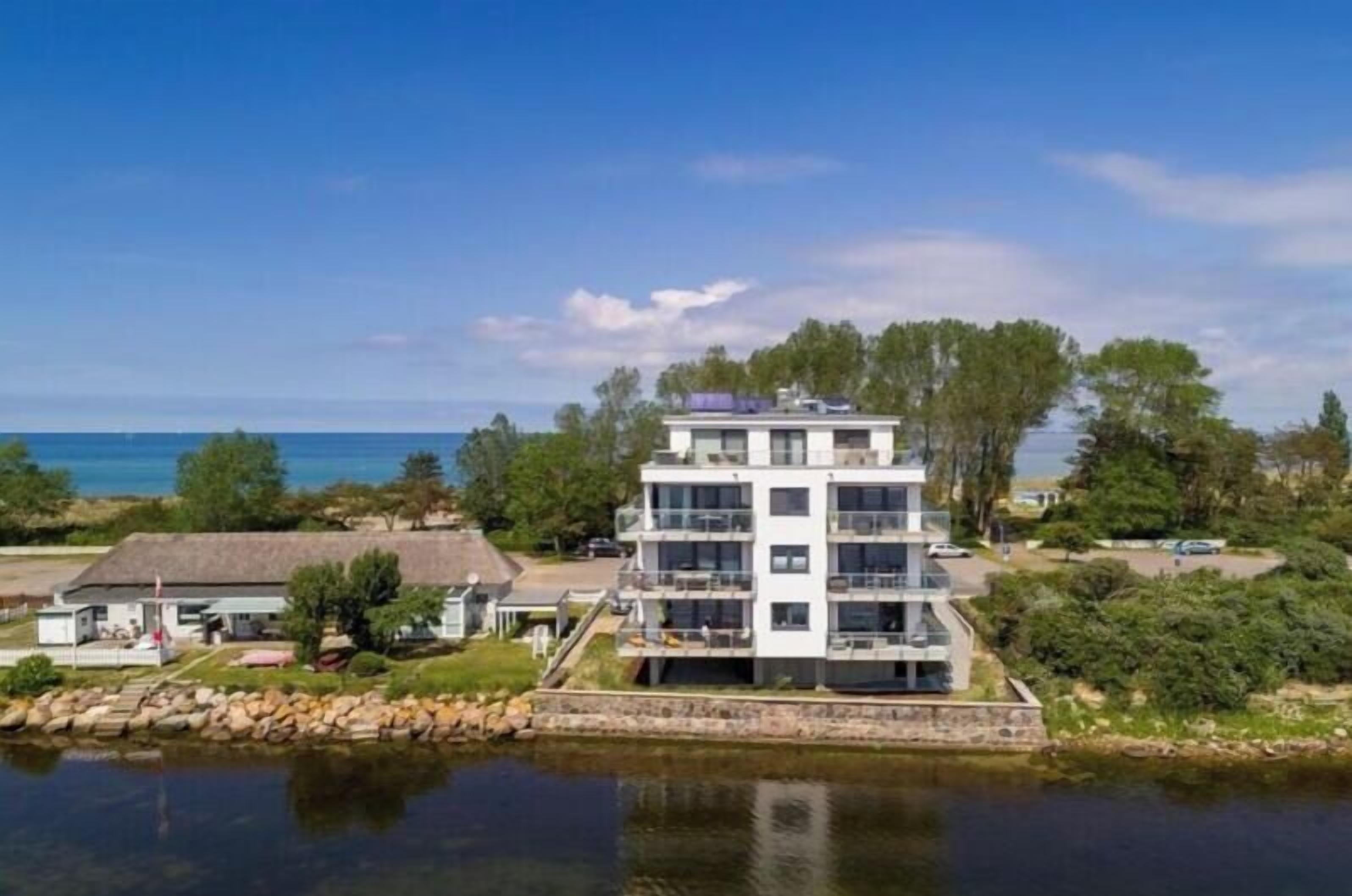 A multi-story building with balconies, situated by a body of water, likely a lake or river, with a clear sky above.