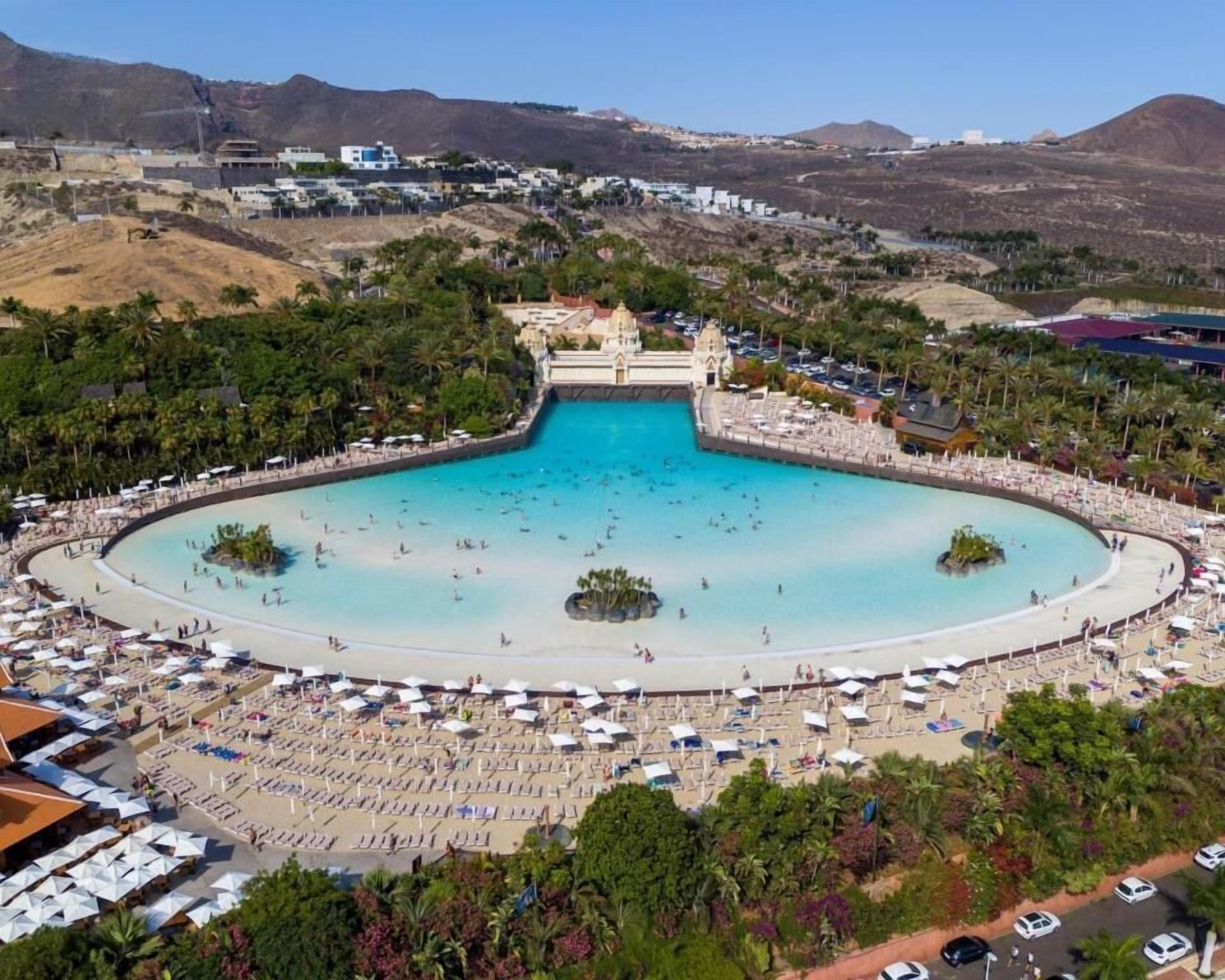 Een luchtbeeld van een resort met een groot centraal zwembad, meerdere strandgebieden en een bergachtige achtergrond.