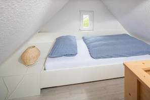Ein kleiner, weißer Raum mit Satteldach, einem Einzelbett, einem kleinen Fenster und einem gewebten Korb.