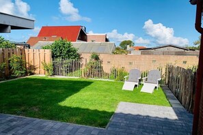 A backyard with a grassy lawn, two white chairs, a wooden fence, and a red-roofed building in the background. - Mariechen am Meer Apt. 2 - Norddeich (Norddeich)