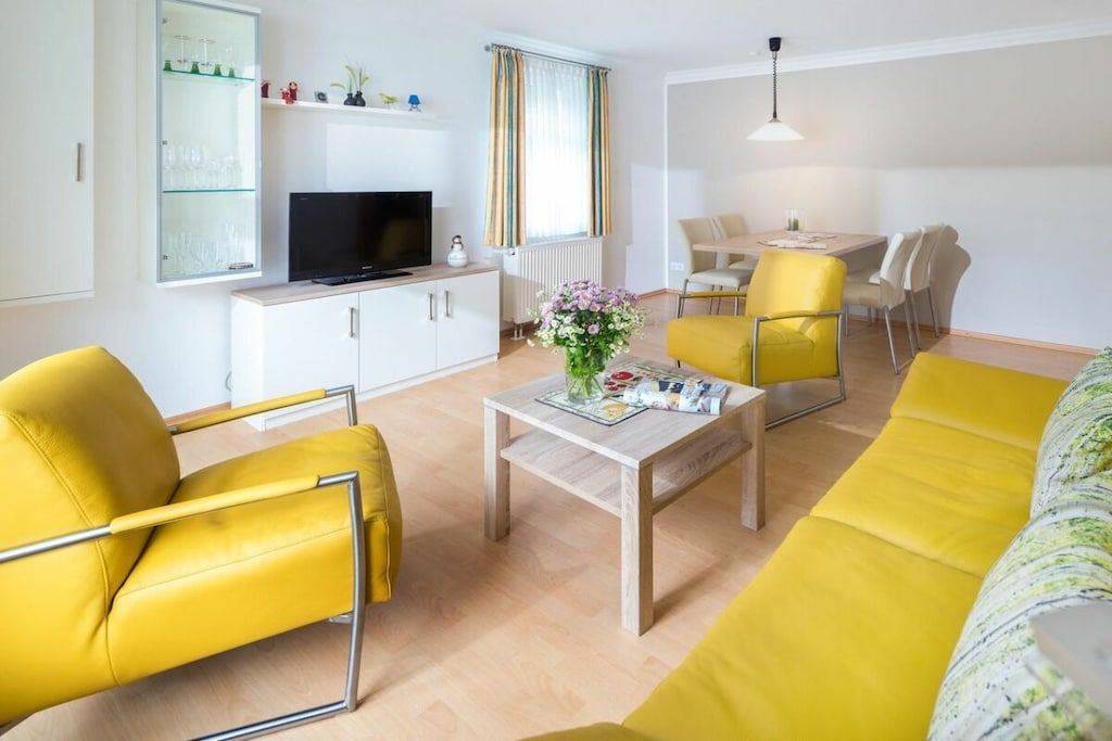 Een moderne woonkamer met geel meubilair, een flatscreen televisie, een eethoek en een raam met gordijnen.